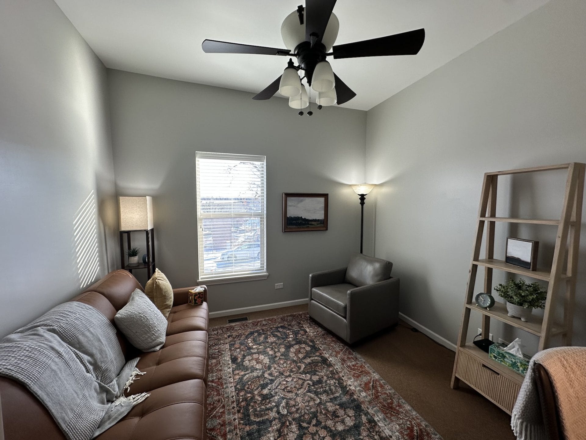 Comfortable counseling office with gray walls, neutral couches and chairs and a book shelf with plants and a clock. 