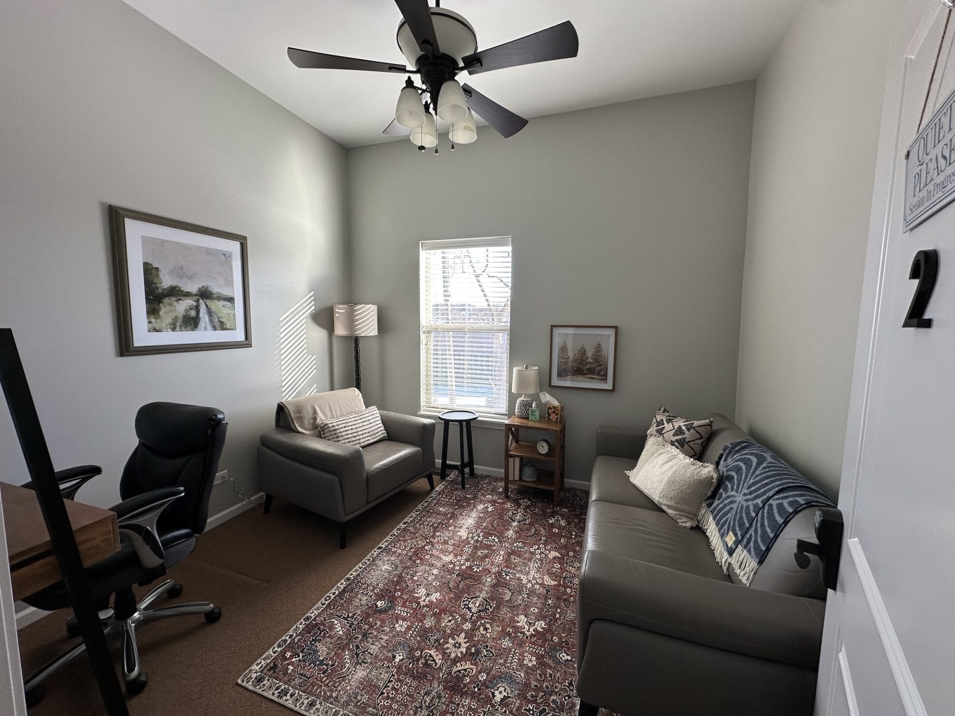 Cozy counseling space with neutral walls, a ceiling fan, art work on the walls and lamps for ambient lighting. 